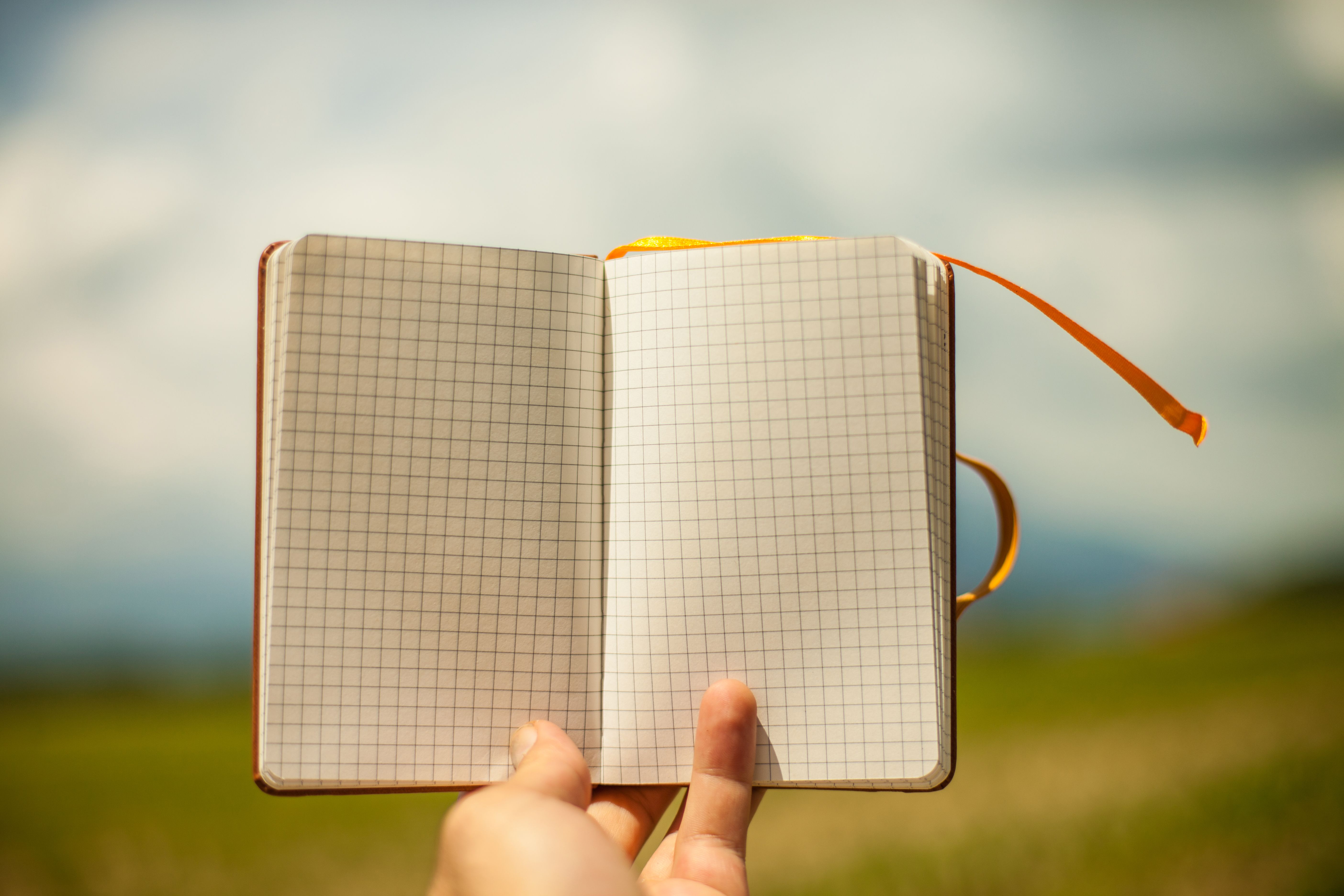Hand holding an open grid notebook with blank pages, outdoors on a sunny day.