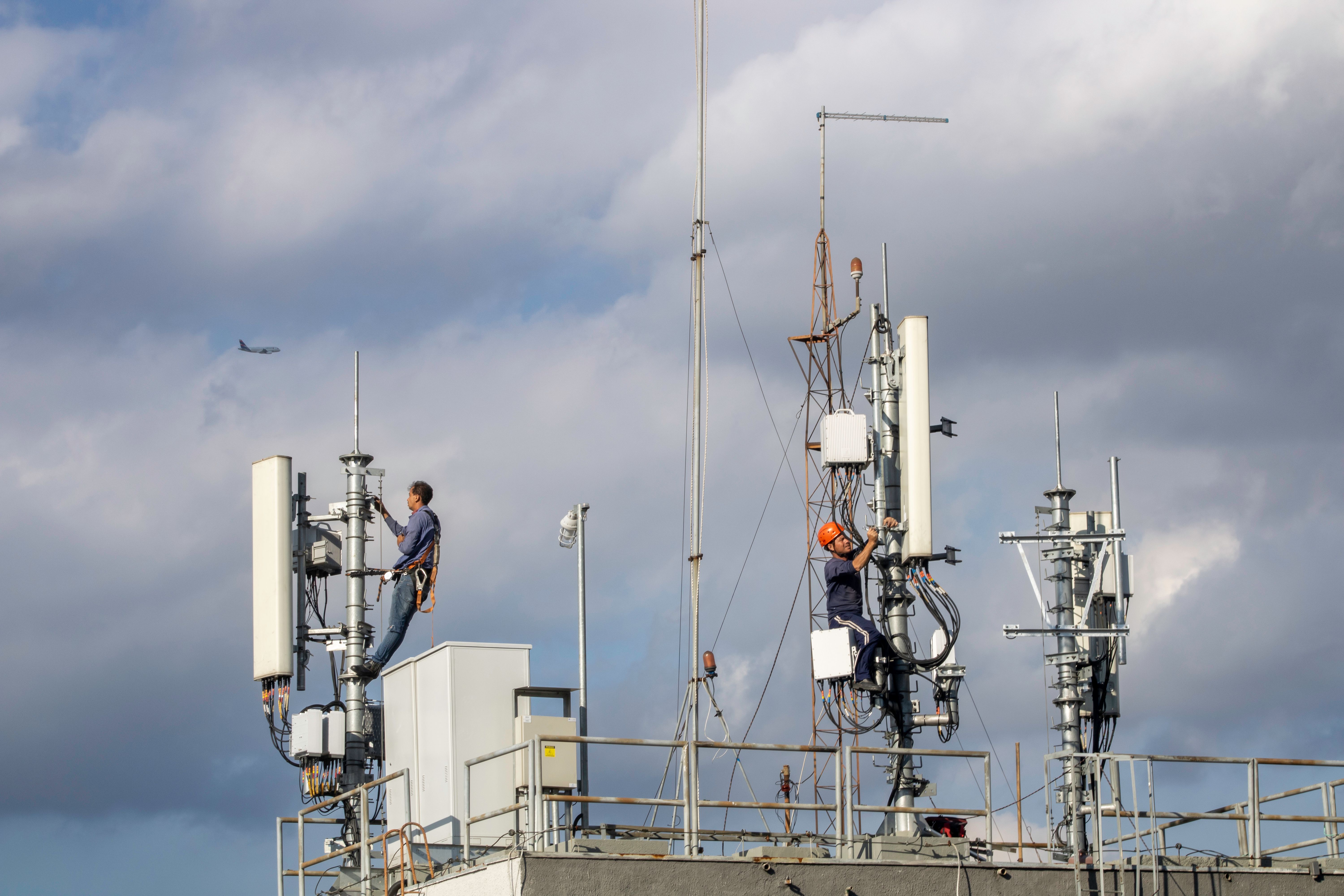 Two workers repairing a cellular tower against a cloudy sky, with an airplane in the background.