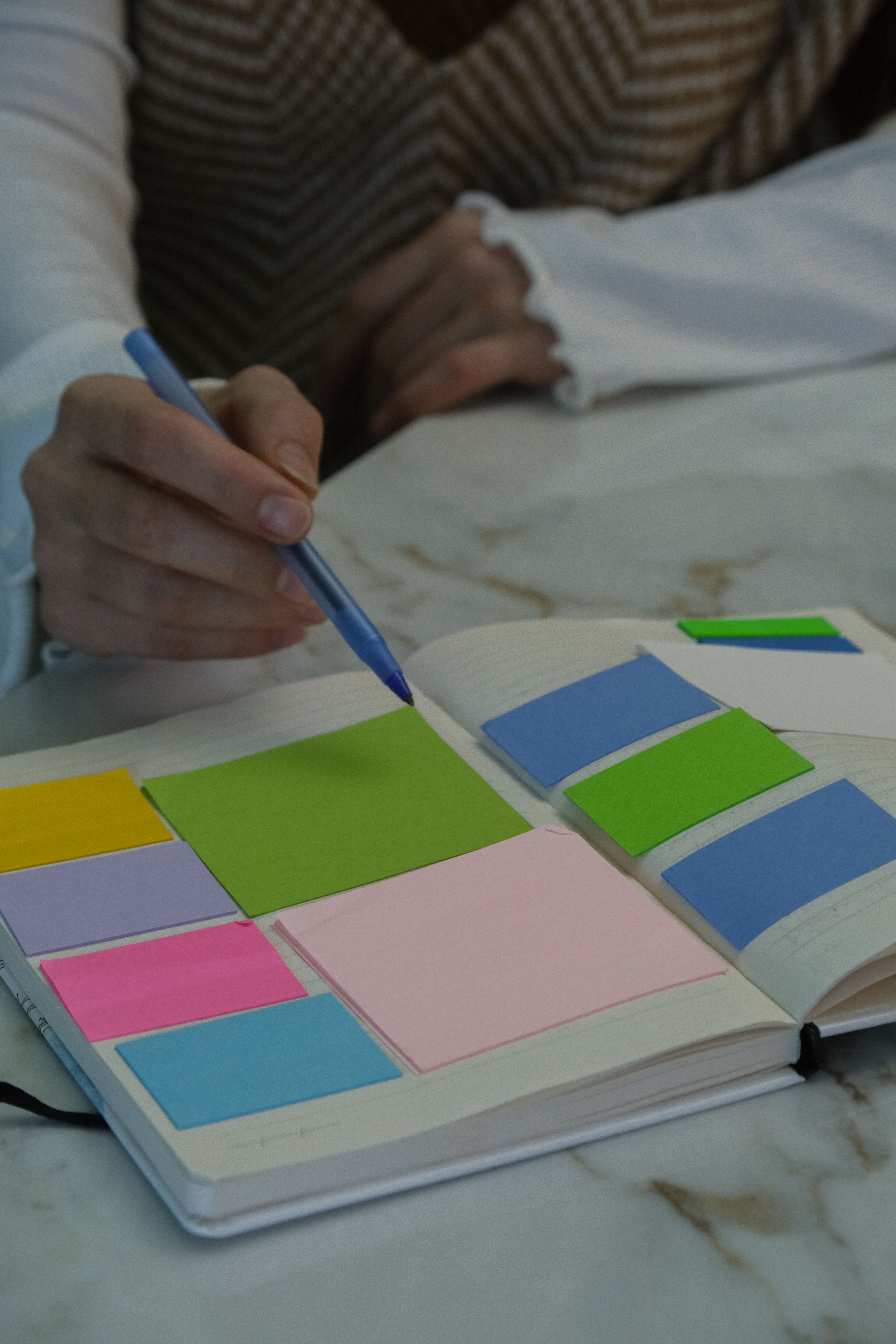 A hand with a pen pointing at colorful sticky notes in a notebook on a marble table.