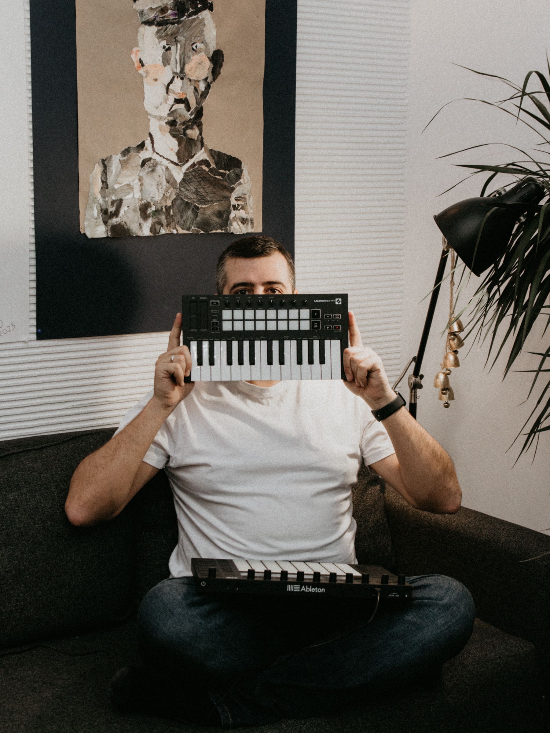 A man in a white t-shirt holds a MIDI keyboard while sitting on a couch in a creative workspace.