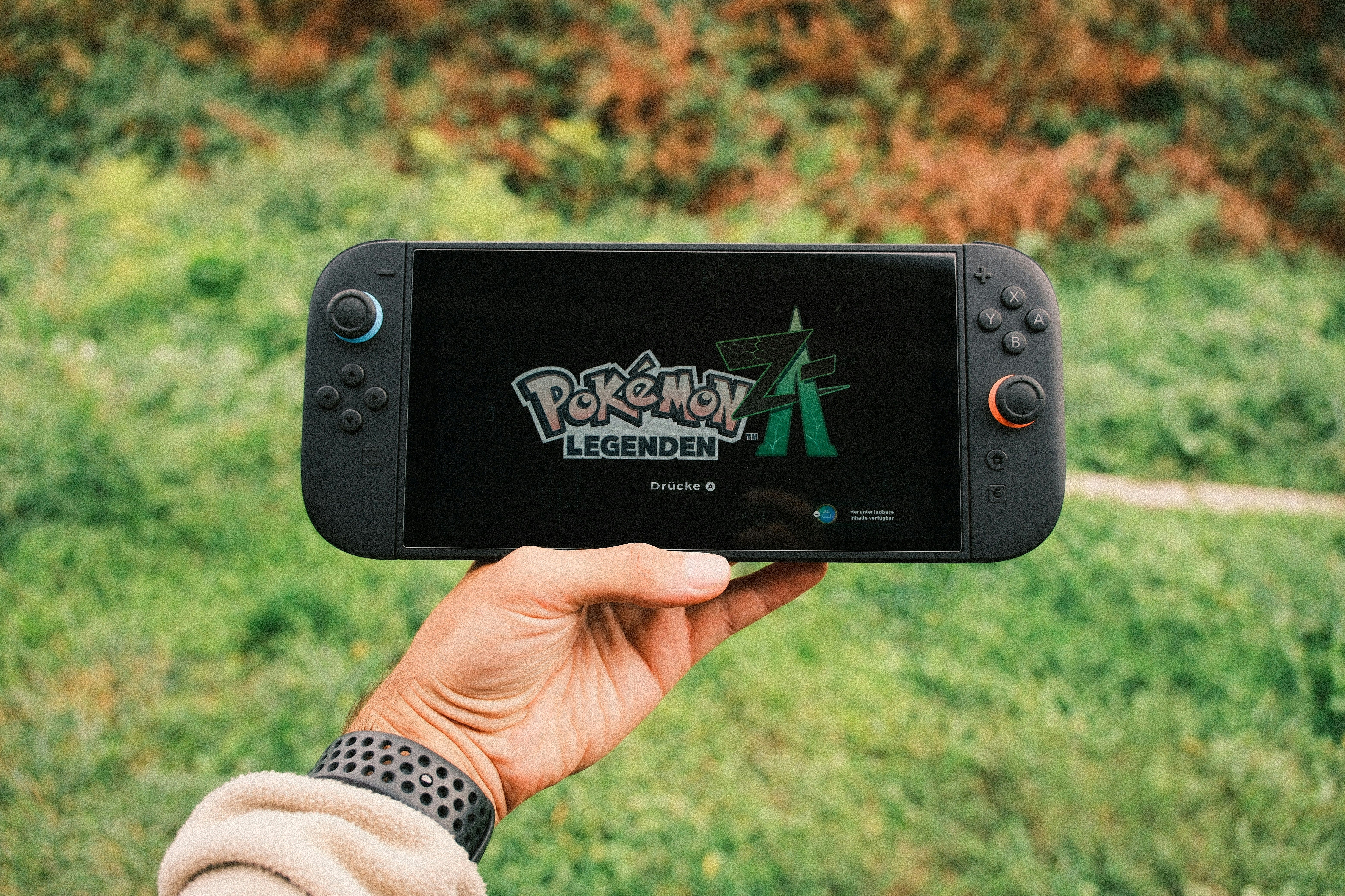 A hand holding a handheld gaming console displaying the Pokémon Legends game screen outdoors.