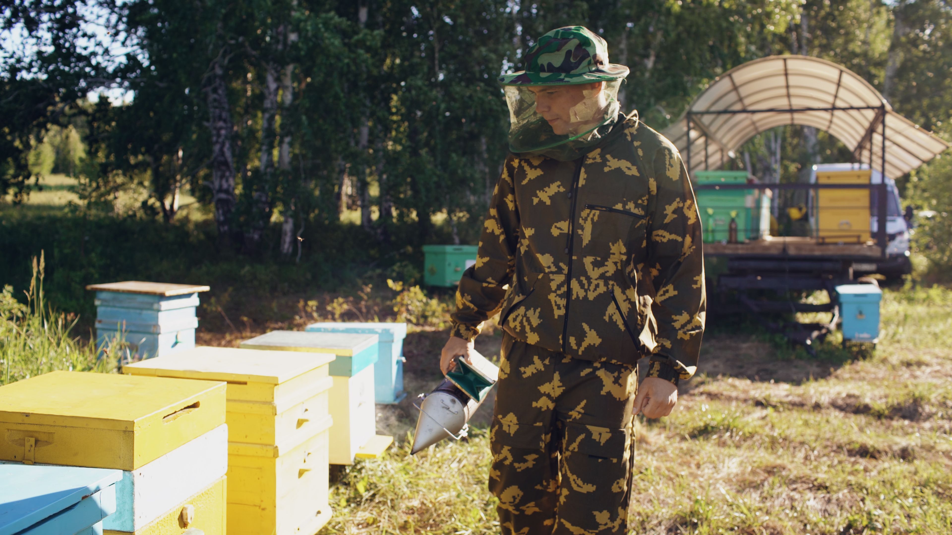 Beekeeper in camouflage suit examining colorful bee hives outdoors.