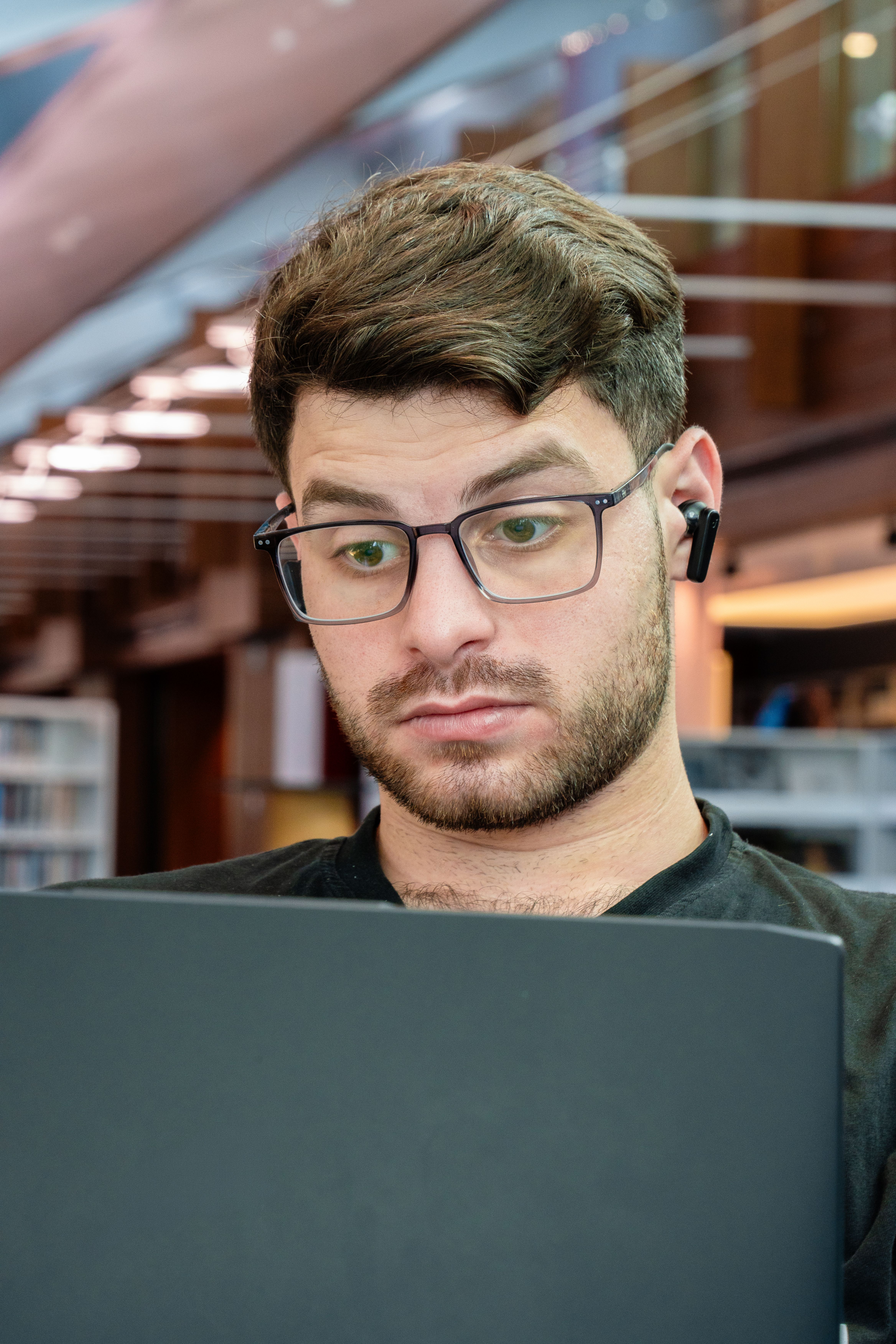 Focused young man with glasses using laptop in a contemporary library setting.
