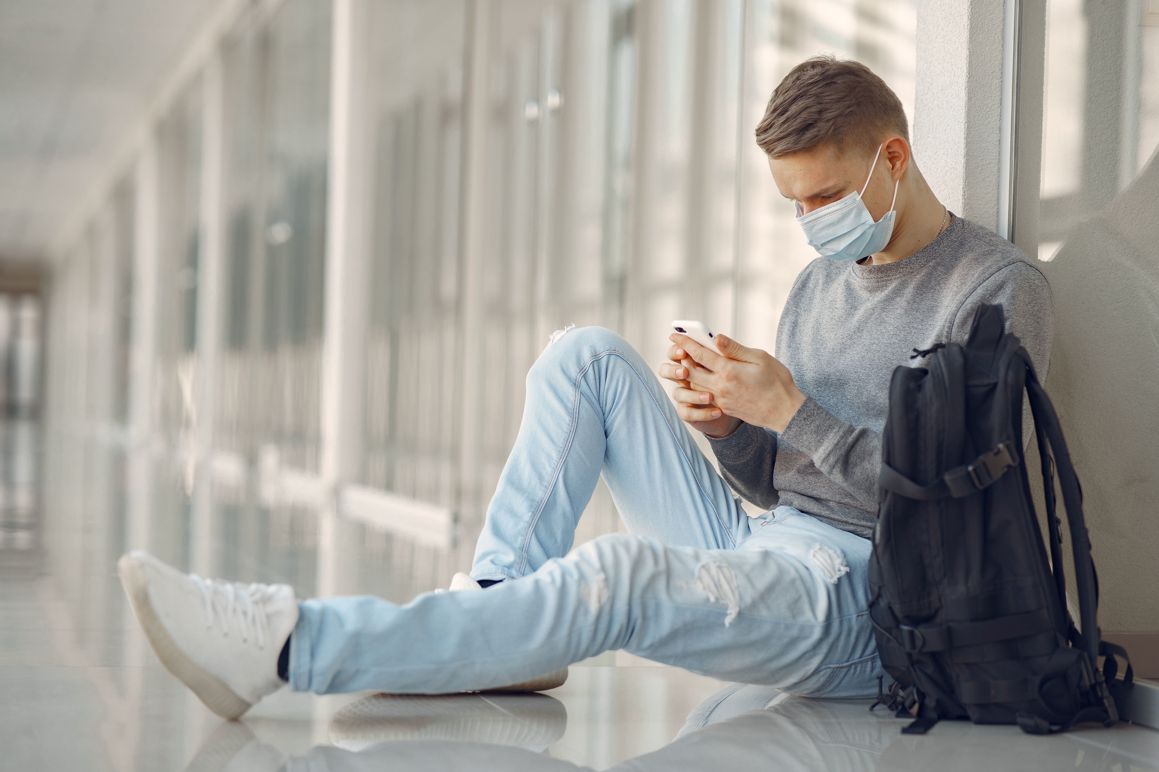 Man in a mask sitting in a hallway, using smartphone, with backpack nearby.