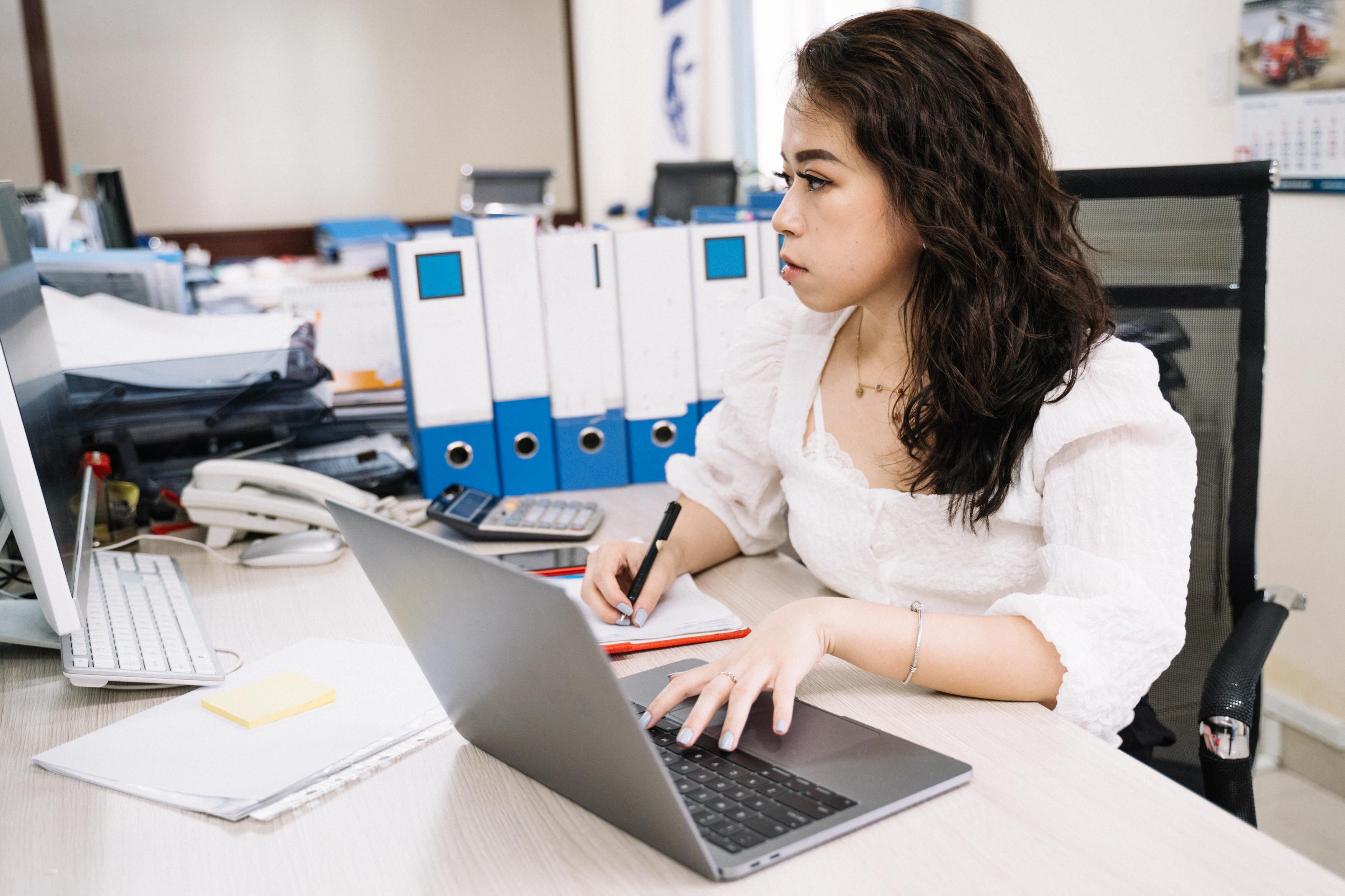 Asian woman attentively working at desk with laptop and paperwork in organized office setting.