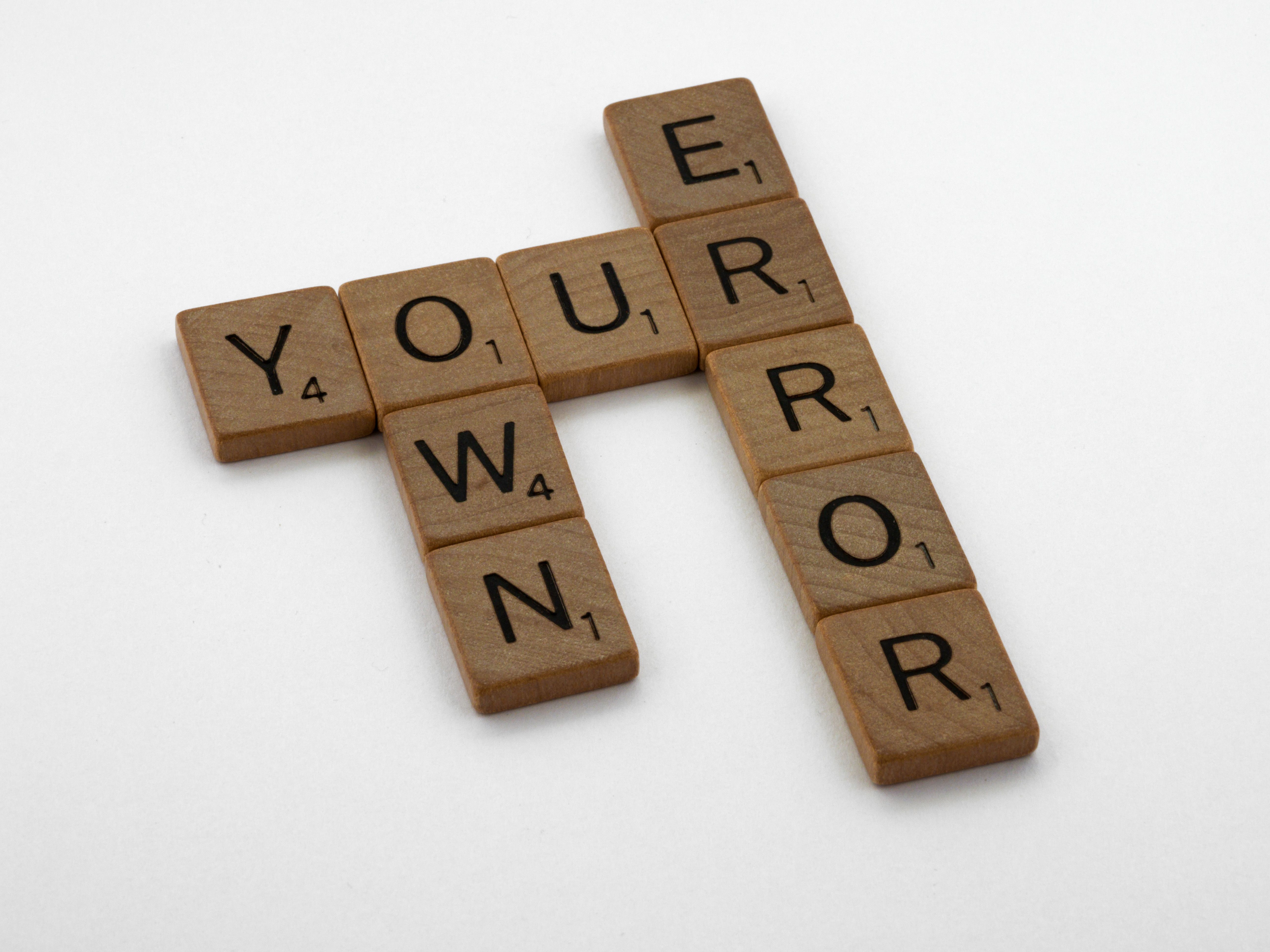 Wooden Scrabble tiles forming the motivational phrase 'Own Your Error' on a white background.