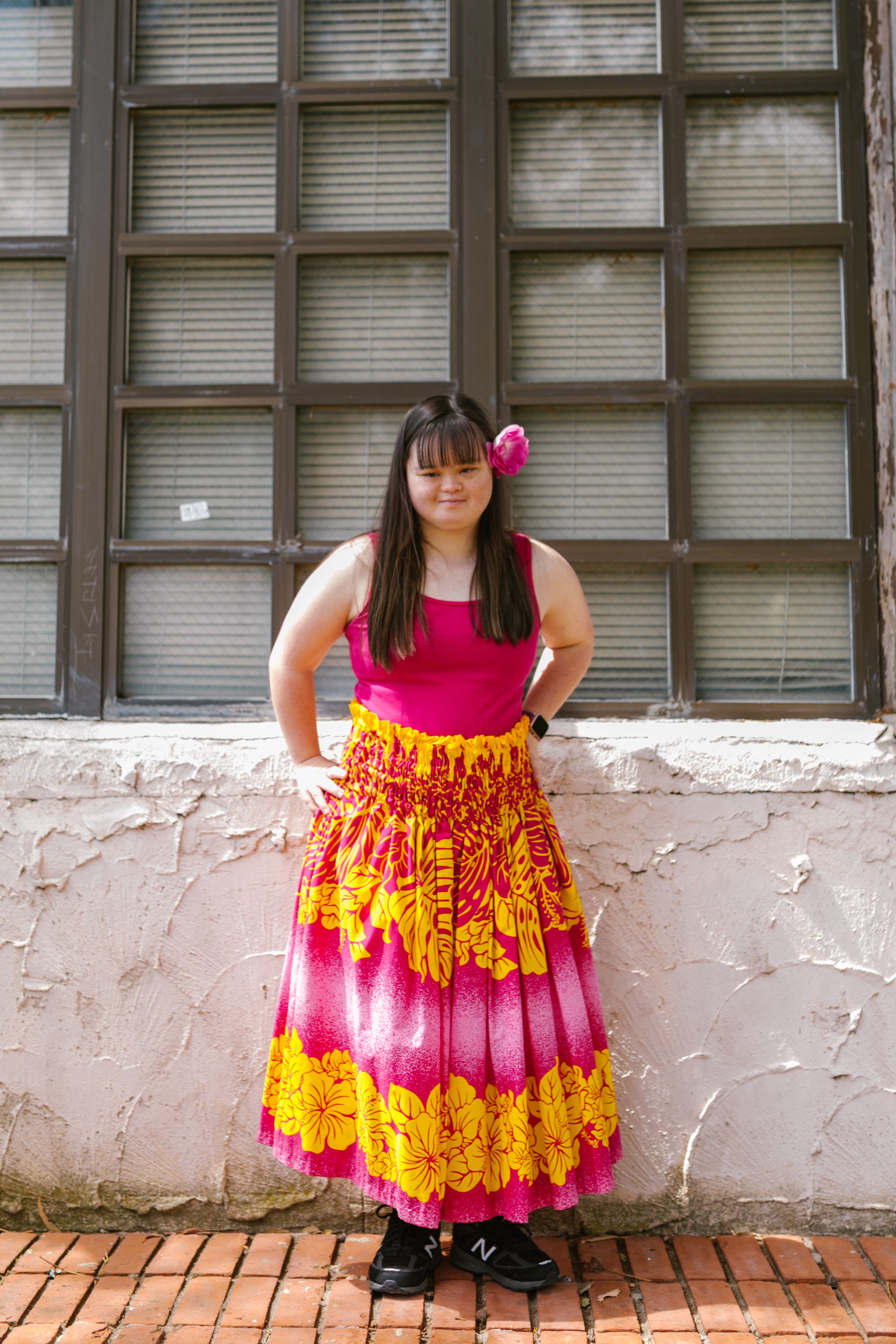 Young woman with Down syndrome poses confidently in a vibrant floral skirt against a windowed wall.
